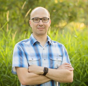 professor wearing a blue and white plaid button up shirt in front of a grassy backdrop
